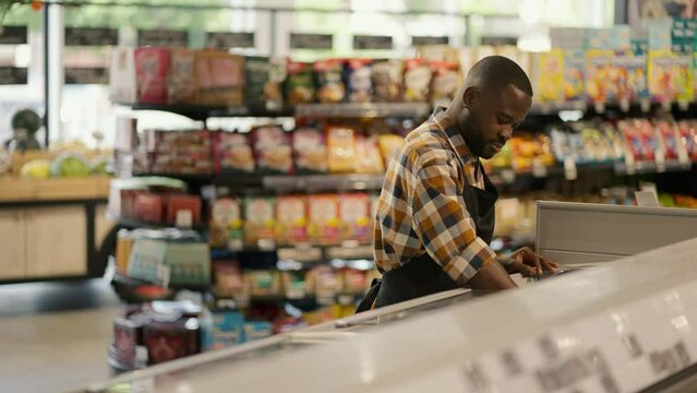 a man with Black skin color in a plaid shirt sorts products in a supermarket refrigerator. Video filmed in high quality