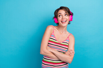 Photo of astonished minded girl folded arms listen music headphones look empty space isolated on blue color background