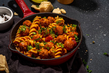 Fusilli pasta with meatballs in tomato vegetable sauce, on dark background with herbs and grated cheese, top view copy space