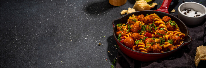 Fusilli pasta with meatballs in tomato vegetable sauce, on dark background with herbs and grated cheese, top view copy space