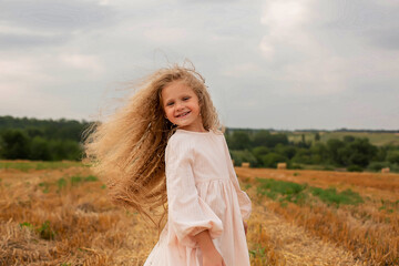 Fototapeta na wymiar a beautiful blonde girl with long hair in a linen dress stands by a haystack in a mown field
