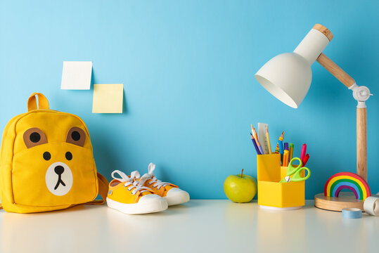 Schooltime treasures: side view photo of desk enriched with school supplies, colorful pencils, ruler, plasticine, shoes, lamp, cute teddy-bear schoolbag and more set against serene blue wall backdrop