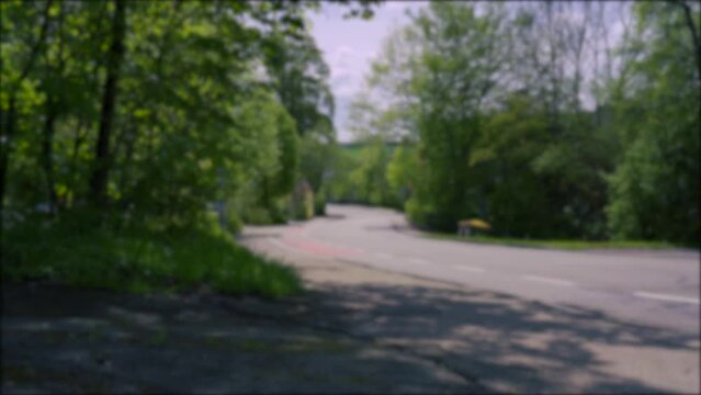 Background, blurry video. A car drives through a quiet green street of a German village among trees.