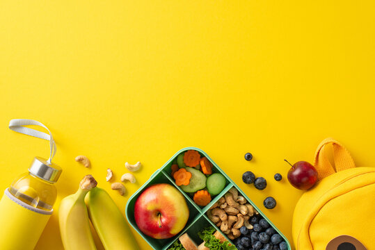 Nourishing school lunch arrangement. Top view showcasing eco-conscious lunchbox with organic treats, water bottle, and playful backpack on yellow background, perfect for advertising or messages