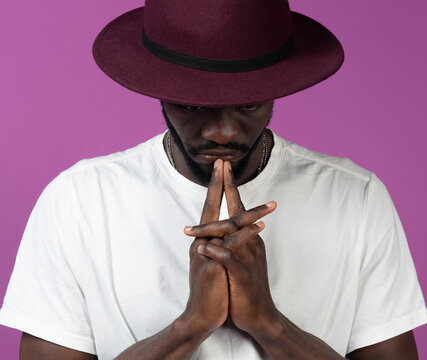 Thoughtful young black man standing in hat near purple wall with fingers near lips