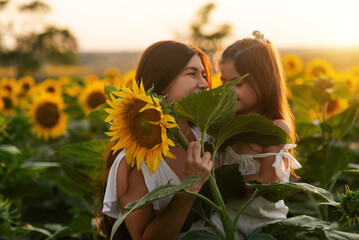 Beautiful young mom cuddling her young daughter among sunflowers at sunset. Motherly love, happy...