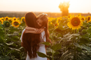 Beautiful young mom cuddling her young daughter among sunflowers at sunset. Motherly love, happy...