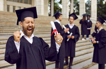 Happy university graduate celebrates graduation. Funny excited overjoyed ecstatic bearded young man in black square student cap and gown standing outside college building holding diploma and screaming