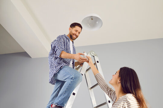 Smiling man changing new electric bulb at home. His wife helping her husband mounting ceiling lamp. Cheerful man standing on ladder holding new LED light bulb, low angle view - Powered by Adobe
