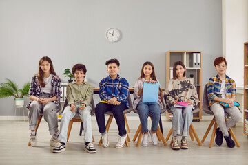 Elementary school students holding notebooks and books while sitting on chairs in row in classroom. Cute boys and girls schoolmates in casual wear listening to teacher during lesson. School education