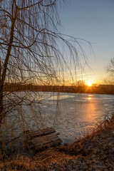 Obraz premium a frozen lake, on the shore of which there are trees and a wooden structure for fishing, against the backdrop of a blue sky with a bright setting sun, during the golden hour