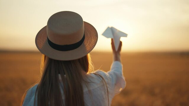 Woman on wheat field holds paper plane in sky at sunset golden hours, back view. Freedom, farm lifestyle, resting in countryside village, outdoor activities concept. Romantic playful female on nature.