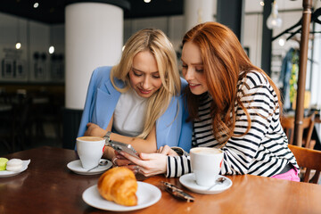 Obraz na płótnie Canvas Portrait of two happy young women sitting at table with coffee at cafe by window, having conversation and showing each other something on mobile phones. Concept of woman friendship