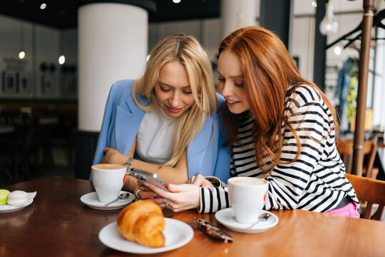 Portrait of two happy young women sitting at table with coffee at cafe by window, having conversation and showing each other something on mobile phones. Concept of woman friendship
