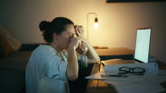 A tired Caucasian woman yawns while sitting in front of her laptop at night working on a project.