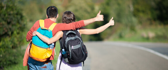 Young couple, backpackers, hitchhiking on a hilly road on a sunny day. Autostop adventure backpack concept. Back view