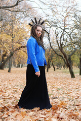 A middle-aged woman in dreadlocks and a hoop on her hair in feathers walks in the autumn park. Woman is looking at camera.