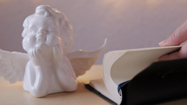 female hand leafs through pages of old black book, family bible in dark cover, white ceramic figurine of angel with wings, concept of intermediary between God and world, all saints' day