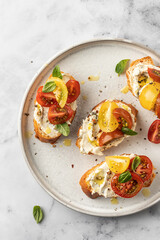 Bruschetta sandwiches with tomatoes, cream cheese, olive oil and basil on a plate on white marble background, top view. Traditional italian antipasti