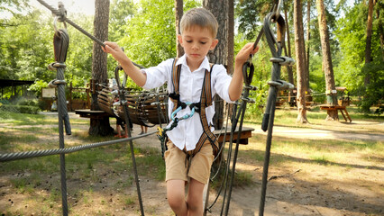 Little boy carefully walking over cable or rope between two trees in park. Active childhood,...