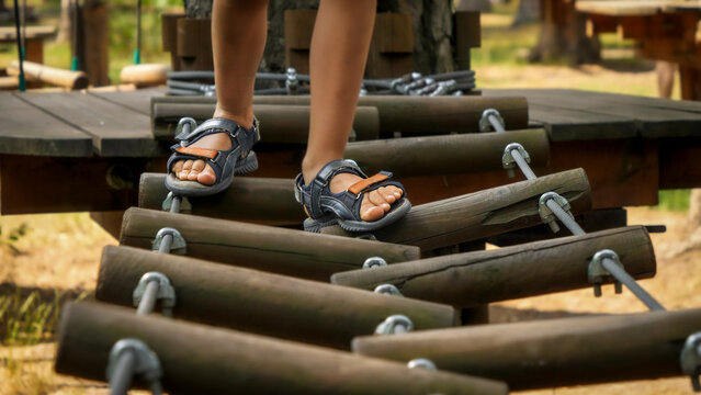 Closeup of childs' feet walking over wobbly rope bridge at adventure park in summer camp. Kids sports, summer holiday, fun outdoors, scouts.
