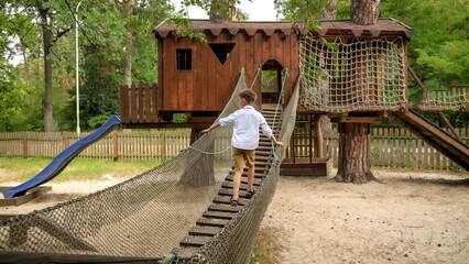 Happy smiling boy walking on the wooden suspension bridge at playground in park. Kids sports, summer holiday, fun outdoors, scouts.