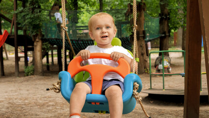 Cheerful toddler boy smiling while swinging in colorful swing on the playground at park