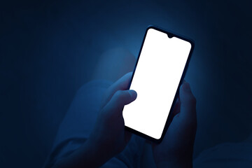 A teenager holds a smartphone with two hands at night. Phone with white screen on defocused dark...