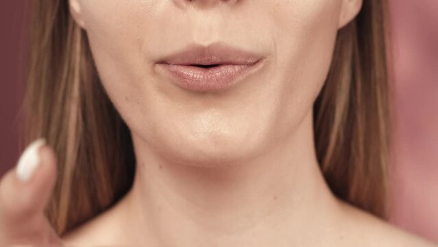 A woman puts her hand with a delicate manicure to her mouth and sends an air kiss. Woman's lips with natural pink gloss close up in the studio on a pink background. Slow motion.