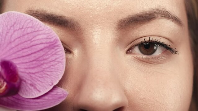 Beautiful eyes of a young woman close up. A woman covers her eye with an orchid flower. Good vision and eye care. Contact lenses. Natural female beauty. Cosmetic line for eye contour care. Slow motion