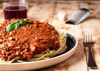Plato de espagueti boloñesa decorado con albahaca sobre un fondo de madera natural junto a un vaso de vino tinto y queso parmesano con un rallador