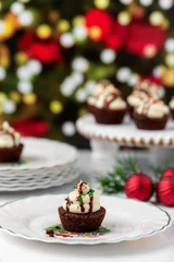 Fototapeten Chocolate Brownie Cupcakes with Buttercream Frosting and Red , Green, and Gold Christmas Sprinkles on White Surface with Christmas Tree in Background  One Cupcake in Front on Christmas Plaates © DiAnna