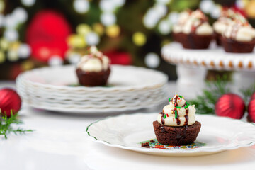 Chocolate Brownie Cupcakes with Buttercream Frosting and Red , Green, and Gold Christmas Sprinkles on White Surface with Christmas Tree in Background; One Cupcake in Front on Christmas Plaates