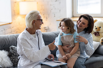 Kind senior female paediatrician doctor examining little child sitting on mother's laps, during home visit or clinic check up. Concept healthcare medical assistance, insurance. Prescribing treatment