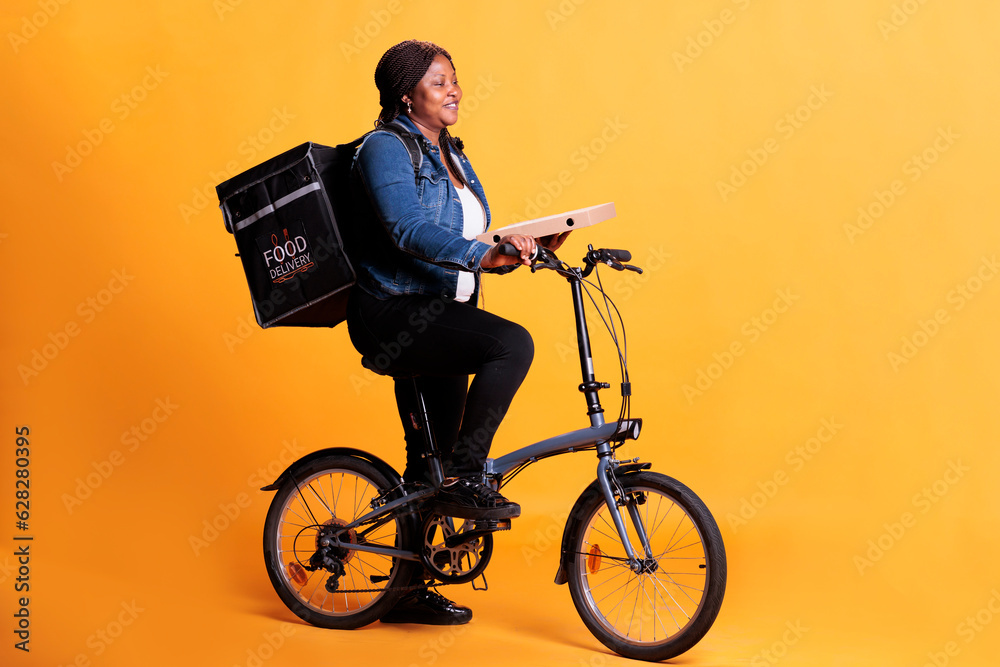 Wall mural African american pizzeria courier carrying pizza carton box delivering takeaway order to client during lunch time, using bike as transportation in studio. Take out food service and concept