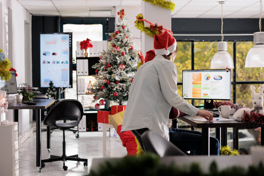 Employees working on business revenue statistical charts in modern office filled with Christmas ornaments. Coworkers wearing holiday season festive clothing doing research for marketing project