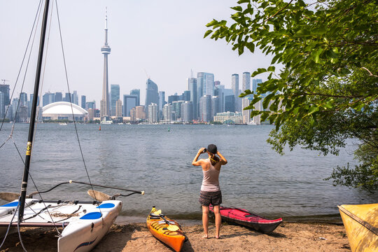 A young woman (back to camera) takes a cell phone picture of the Toronto city skyline from a beach in the Inner harbour with kayaks and a sailboat in the fore ground on a hot summer aftertnoon