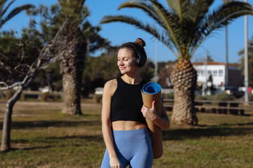 Young sporty woman preparing for training, yoga practice, pilates outdoors. Girl in headphones and sportswear with mat working out on summer vacation at park with palm trees. Sports, healthy lifestyle