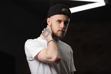 Stylish bearded caucasian young man with tattooed arm in cap and white t shirt looking at camera,...