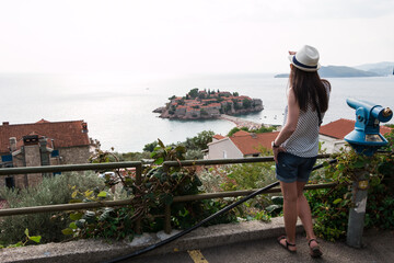 girl tourist in a hat looks from a top on the island of Sveti Stefan in Montenegro and the sea