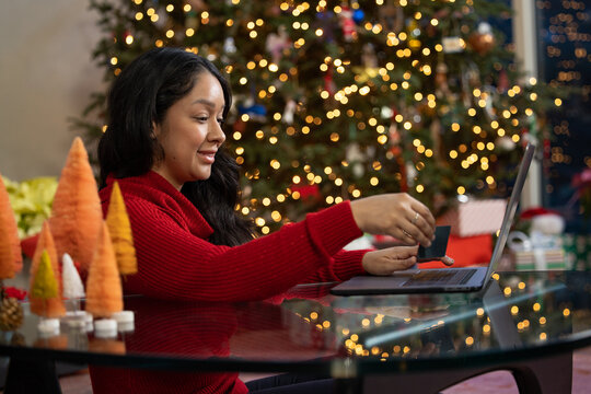 Hispanic woman holding credit card in her right hand while online shopping on her computer in front of her Christmas tree at home
