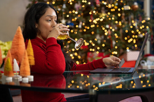 A hispanic woman taking a drink of wine while online shopping on her computer in front of her Christmas tree at home
