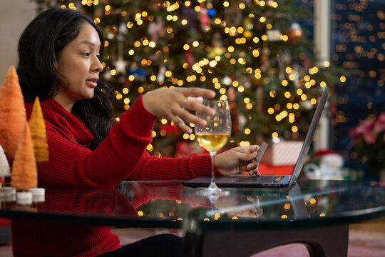 Hispanic woman reaching for her glass of wine while online shopping on her computer in front of her Christmas tree at home