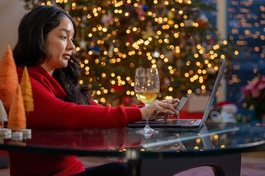 Hispanic Woman Holding A Credit Card Shopping Online On Her Computer With A Glass Of Wine Beside In Front Of Her Christmas Tree At Home