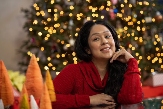 Hispanic woman with her left hand under her chin in front of her Christmas tree at home