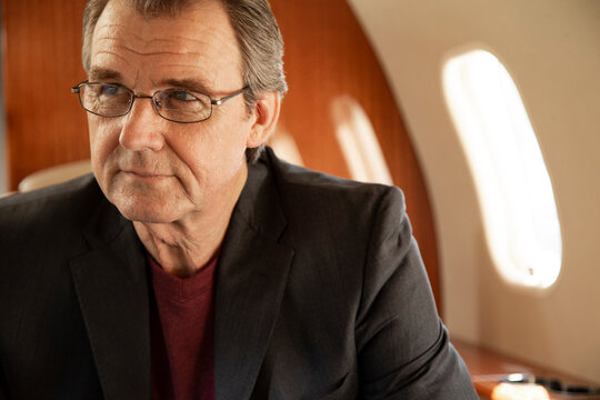 Portrait of mature business man wearing glasses sitting in seat aboard private jet looking off camera 