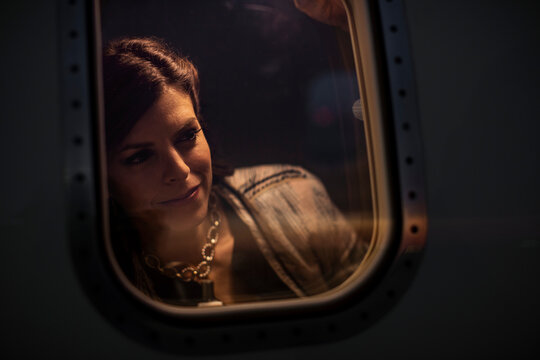 Middle aged woman looking out of window of private jet admiring the sunset, view from outside of plane 