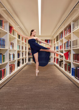 Young female ballerina striking pose in library pulling one book out with her hand and pushing another in with her foot 