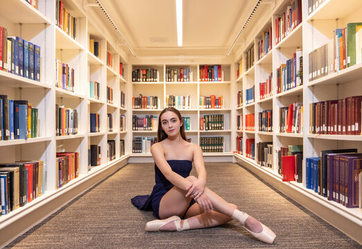 Fototapeta Portrait of young female ballerina sitting on floor of library pulling looking to camera 