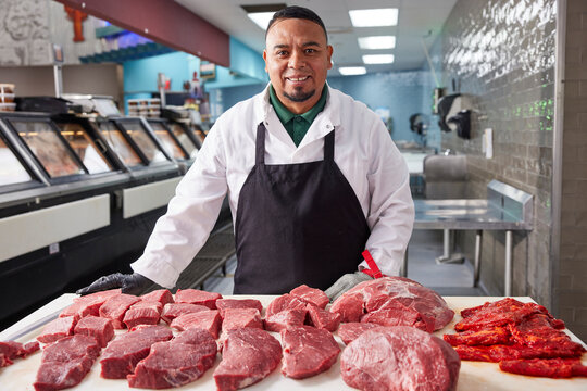 Smiling portrait of butcher wearing hair net behind counter of meat market of grocery store with selection of cuts of steak 